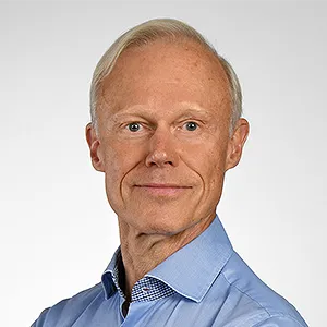 ein älterer Mann mit grauen Haaren und hoher Stirn. Er trägt ein blaues Hemd und lächelt