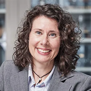 Portrait einer Frau mit dunkelbraunen gelocketen Haaren. Sie trägt einen grauen Blazer und lächelt in die Kamera